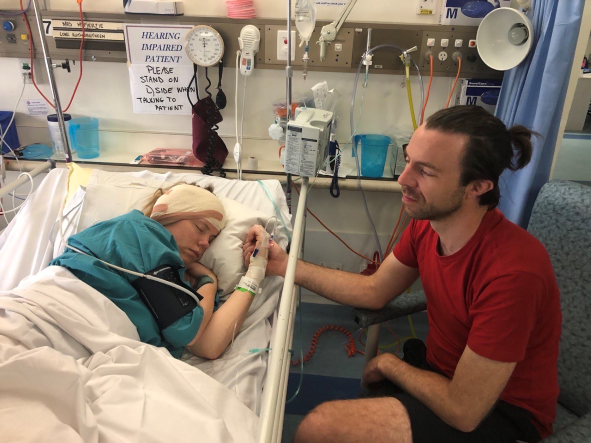 A young woman is lying in her hospital bed asleep with a bandage on her head, her husband sits next to her holding her hand.